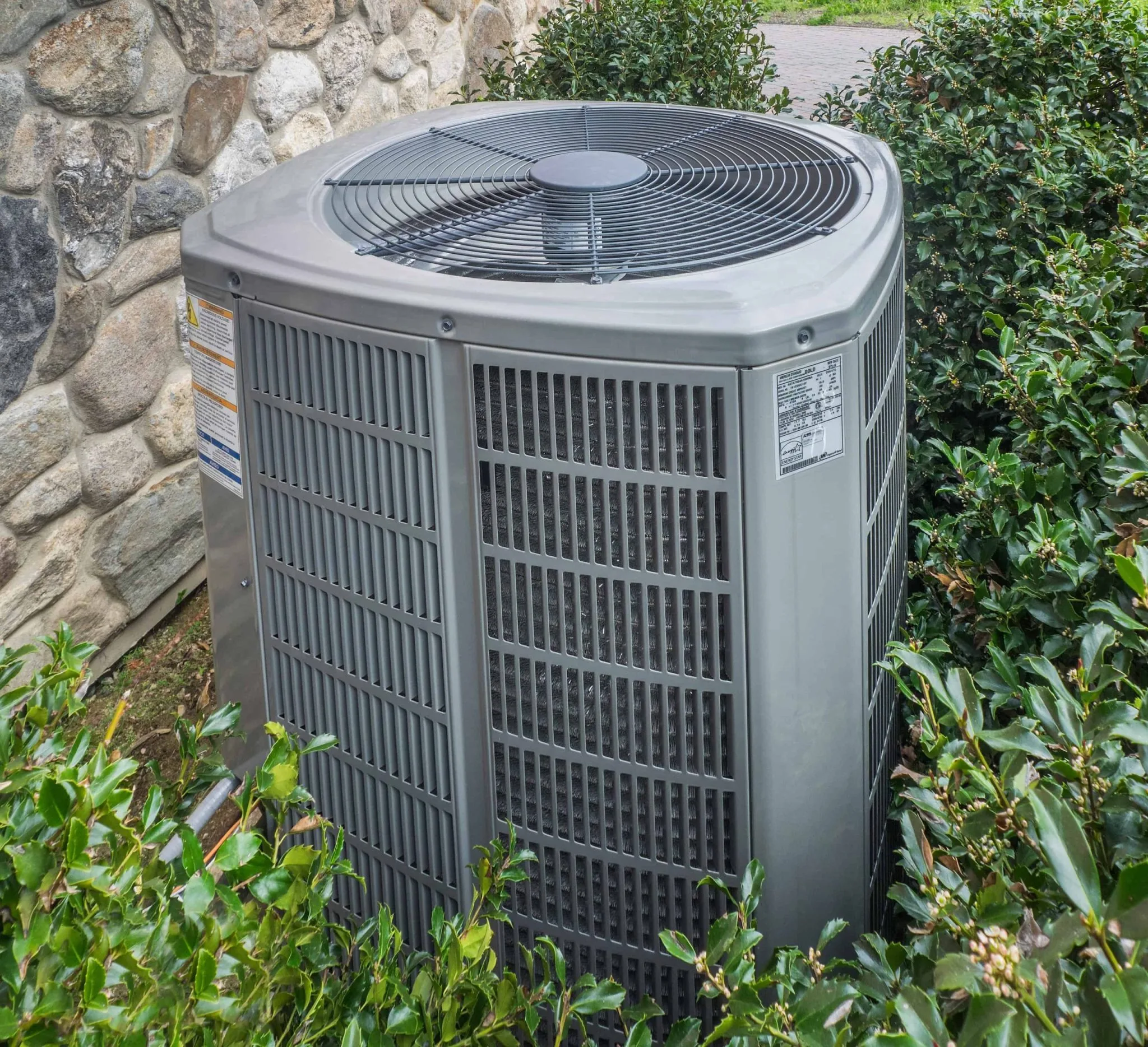 A central air conditioning unit sits outside next to a stone wall in Queens, NY, surrounded by green bushes. The metal unit features a large fan on top and vented sides for airflow—ideal for homes needing emergency HVAC service Long Island.