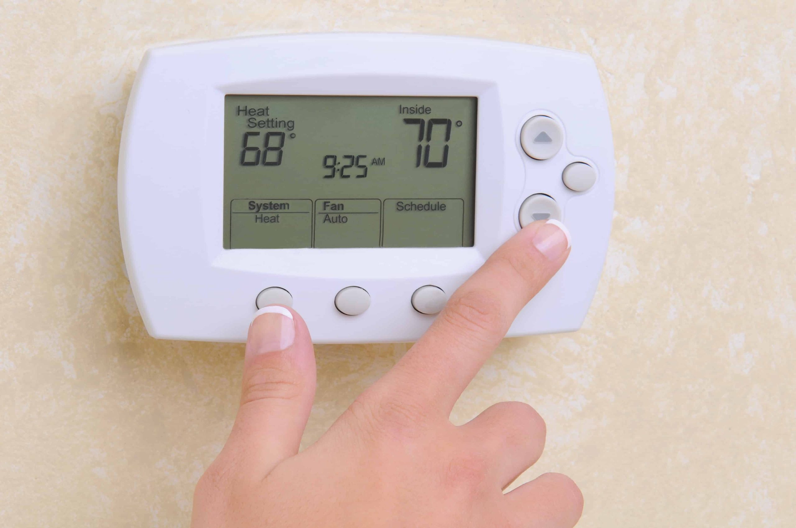 A hand presses a button on a digital thermostat mounted on a beige wall. The display shows 68°F heat, 70°F inside temperature, and 9:25 AM—perfect for homes in Queens, NY needing emergency HVAC service on Long Island.