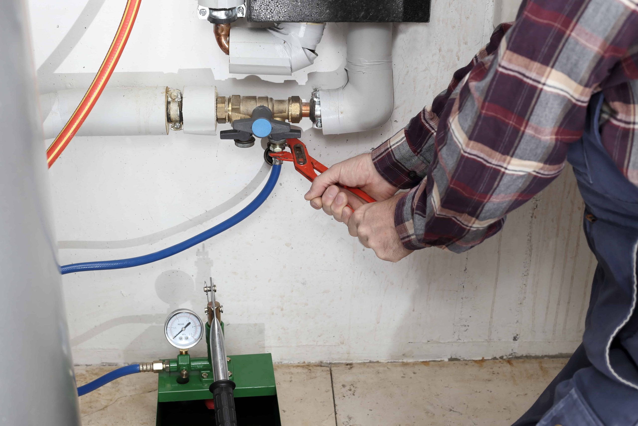 A person in a plaid shirt uses a red pipe wrench to tighten a valve on a plumbing system with pipes and a pressure gauge, providing emergency HVAC service Long Island style, all against a white wall.