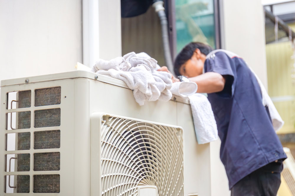 A person in a dark uniform services an outdoor air conditioning unit near a building wall in Queens, NY. Several white towels rest on top of the unit, and the individual’s face is not visible—likely providing emergency HVAC service Long Island needs.