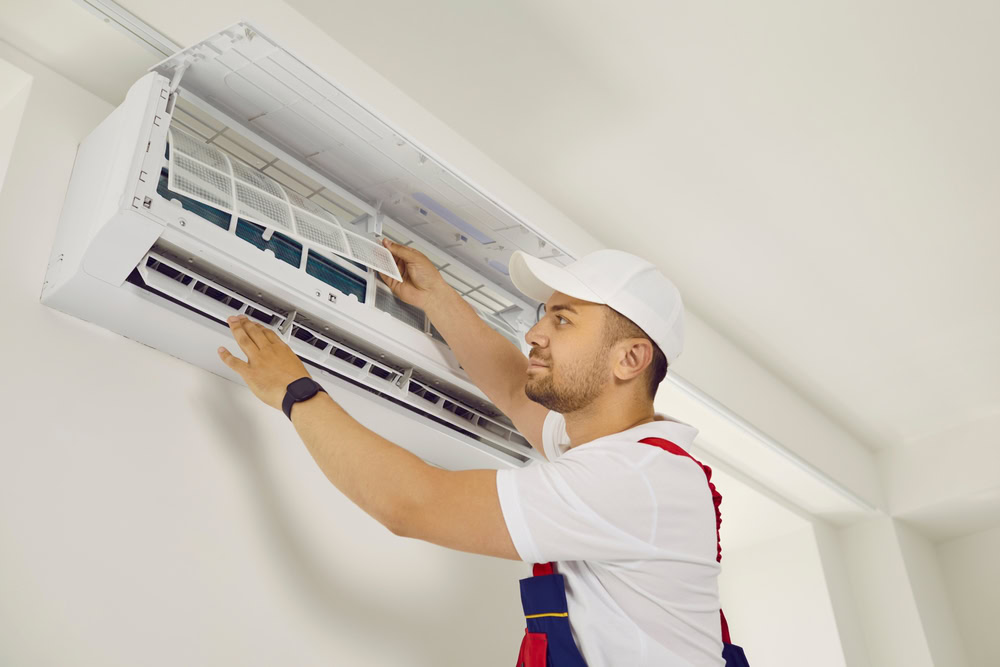 A technician wearing a white cap and uniform is inspecting or repairing an air conditioning unit mounted on a wall in Queens, NY. He is lifting the front cover, checking the filters inside—ready to provide emergency HVAC service Long Island residents trust.