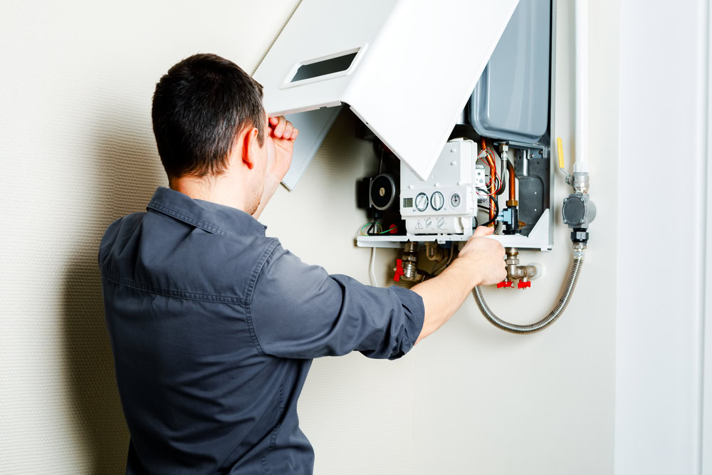 A man in a gray shirt repairs or inspects a wall-mounted gas boiler in Queens, NY, with the cover lifted open to reveal internal components—ideal for emergency HVAC service Long Island needs.