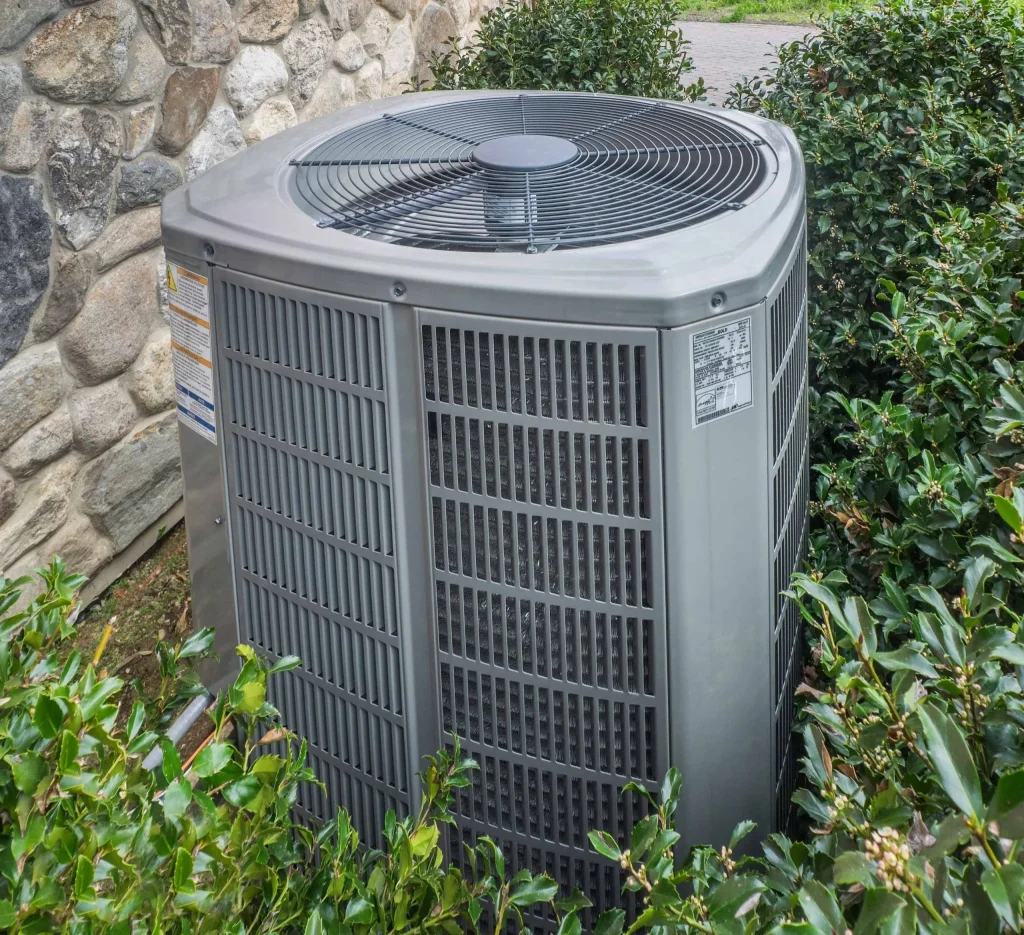 A central air conditioning unit sits outdoors next to a stone wall in Queens, NY, surrounded by green bushes and plants. The gray metal unit has ventilation grilles and a large fan on top—perfect for homes needing emergency HVAC service on Long Island.
