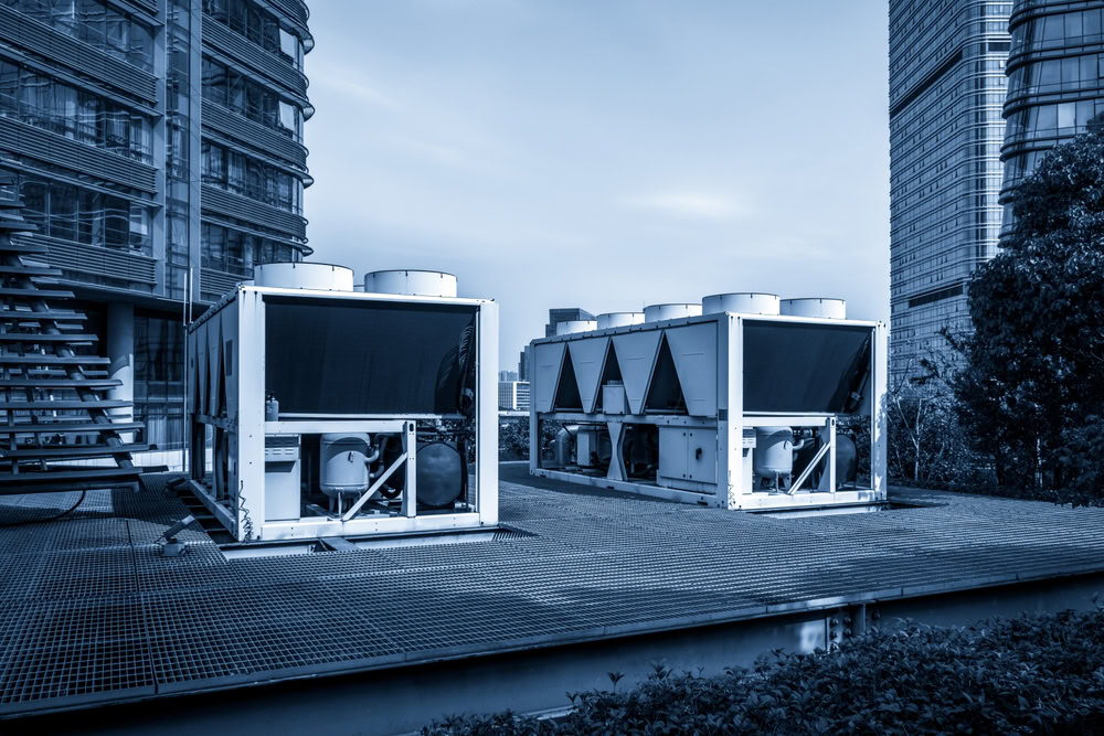 Two large industrial HVAC units are installed on a metal rooftop platform in Queens, NY, with modern office buildings and greenery visible in the background under a blue-toned sky. Call for emergency HVAC service Long Island if needed.