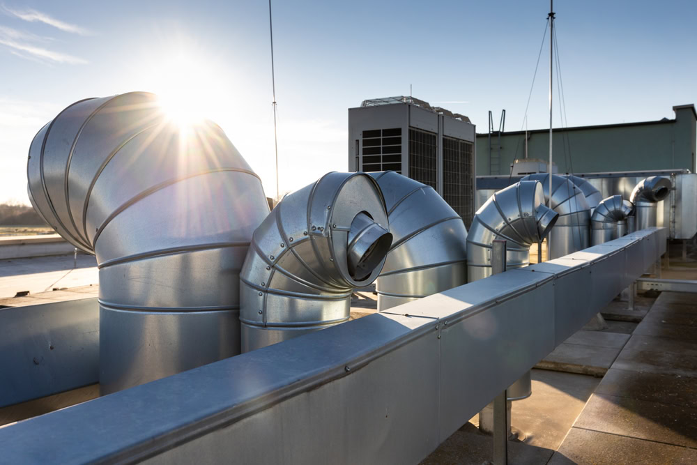 Large metal ventilation ducts and pipes on a Queens rooftop in NY shine under sunlight, with an HVAC unit nearby, ready for emergency HVAC service Long Island, all set against a clear blue sky.