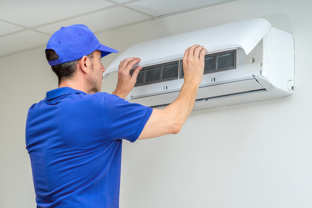 A technician in a blue uniform and cap is inspecting or repairing a wall-mounted air conditioner with the front cover open, providing emergency HVAC service in Long Island, NY.