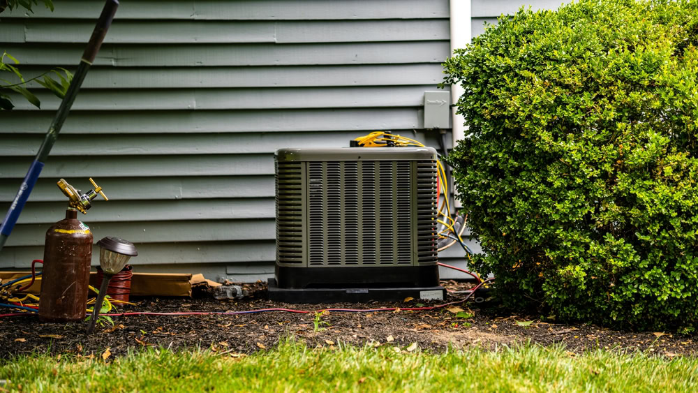 An air conditioning unit is installed outside a house in Queens, NY, next to a large green bush and gray siding. Nearby, HVAC service tools and a small gas tank are placed on the ground for emergency HVAC service Long Island professionals.