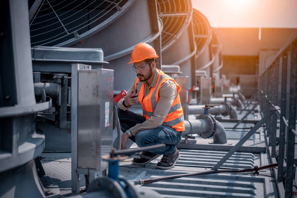 A construction worker in a safety vest and hard hat kneels beside industrial HVAC equipment in Queens, NY, inspecting a control panel and talking on a walkie-talkie—ready to provide emergency HVAC service when needed. Large fans and pipes fill the background.