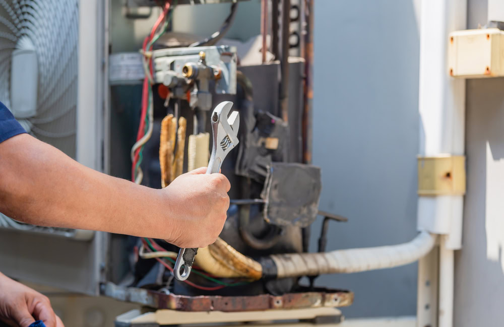 A person’s hand holds two wrenches in front of an open air conditioning unit with exposed wires and pipes, performing maintenance or emergency HVAC service outdoors in Queens, NY.