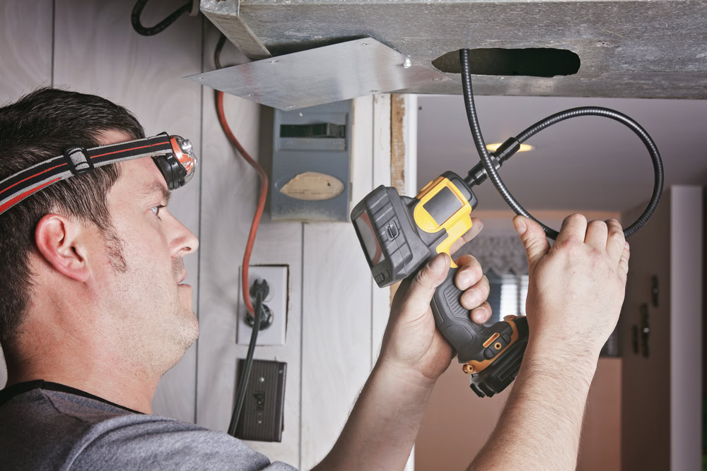 A man wearing a headlamp uses an inspection camera to look into an air duct or vent, inspecting or repairing part of an HVAC system in a Queens, NY residential setting.