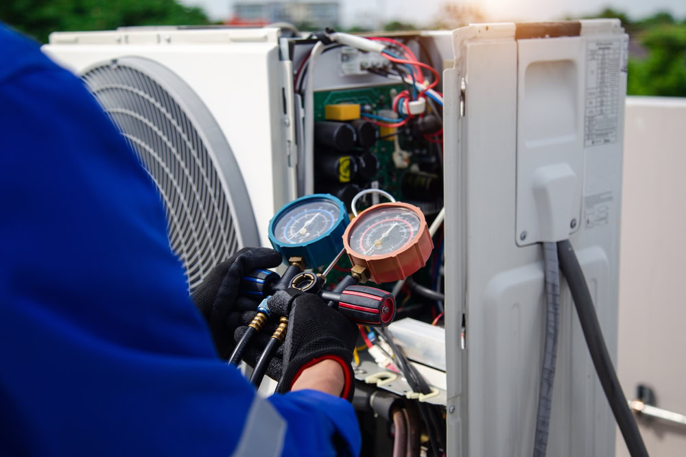A technician in blue uniform and black gloves uses diagnostic gauges to check or repair an outdoor air conditioning unit in Queens, NY, with the unit’s panel open exposing internal components—ideal for emergency HVAC service Long Island needs.