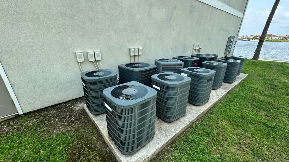 Ten outdoor air conditioning units are lined up on a concrete pad beside a building in Long Island, NY. Grass and a lake are nearby, with electrical boxes above. For fast repairs, emergency HVAC service in Long Island, NY and Queens is available.