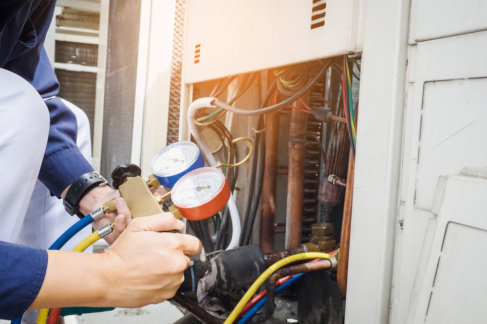 A technician in Queens uses a manifold gauge to check the pressure of an air conditioning unit, with colorful hoses and exposed wires visible inside the open panel—providing emergency HVAC service Long Island residents can trust.