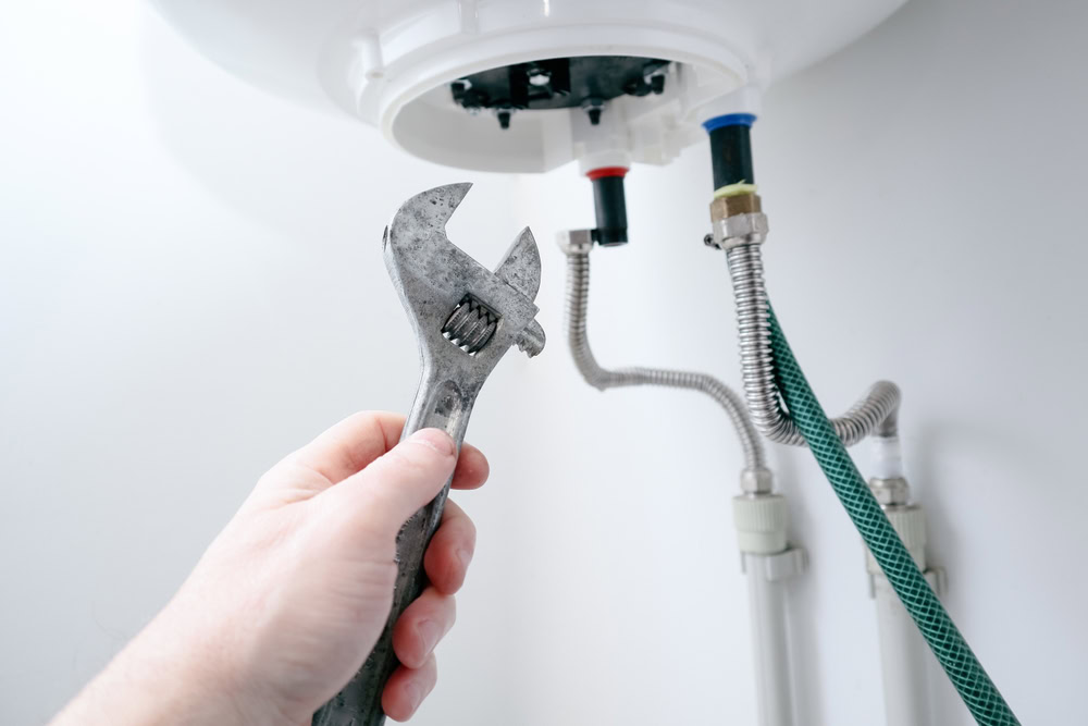 A hand holding a wrench is positioned near pipes and fittings under a water heater in Queens, NY, appearing ready to perform maintenance or repair—ideal for emergency HVAC service Long Island residents may need.