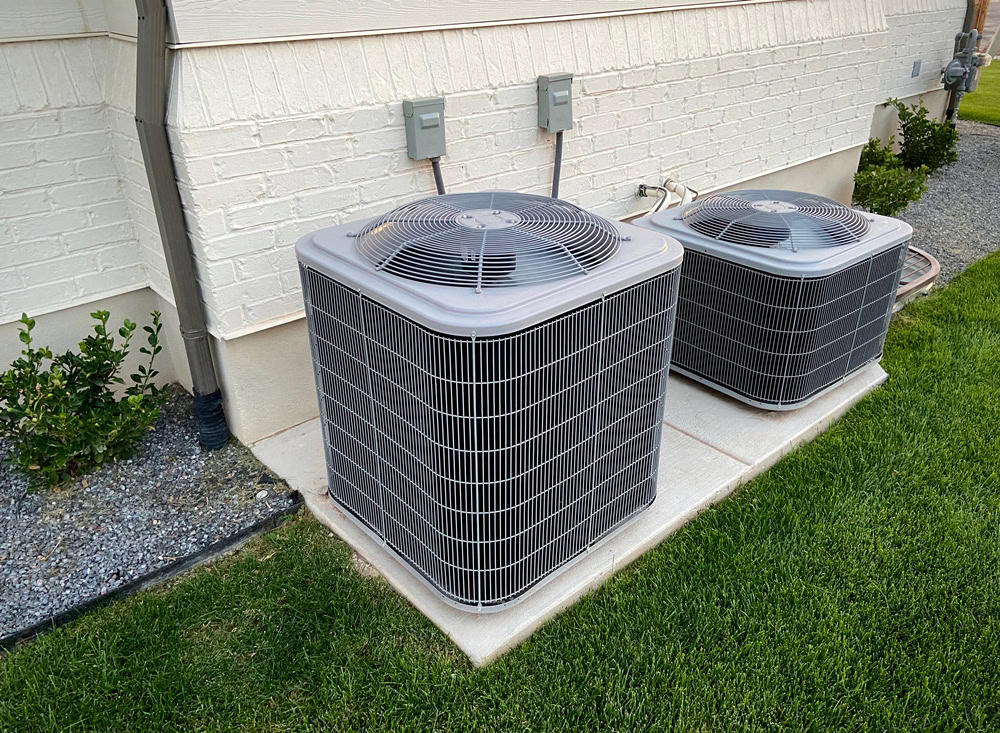 Two outdoor air conditioning units sit on concrete pads next to a house in Queens, NY, surrounded by green grass and small shrubs, with electrical boxes mounted on the wall above them—ideal for emergency HVAC service Long Island residents may need.
