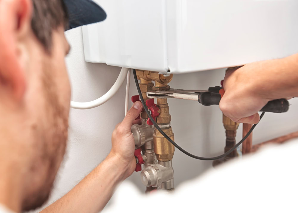 A plumber in Queens uses pliers to adjust pipes and valves under a wall-mounted boiler, performing maintenance or installation—ready to respond with emergency HVAC service Long Island residents can rely on.