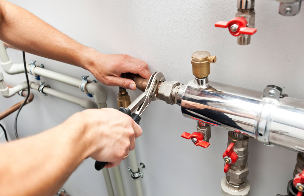 A person uses a wrench to tighten a pipe fitting on a shiny metal plumbing system with multiple valves and connected pipes against a white wall, similar to those serviced by emergency HVAC service Long Island professionals.
