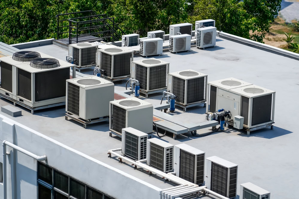 Multiple air conditioning units and HVAC systems are installed on a flat rooftop, surrounded by trees and greenery in the background, illustrating the type of setup serviced by emergency HVAC service Long Island and Queens, NY professionals.