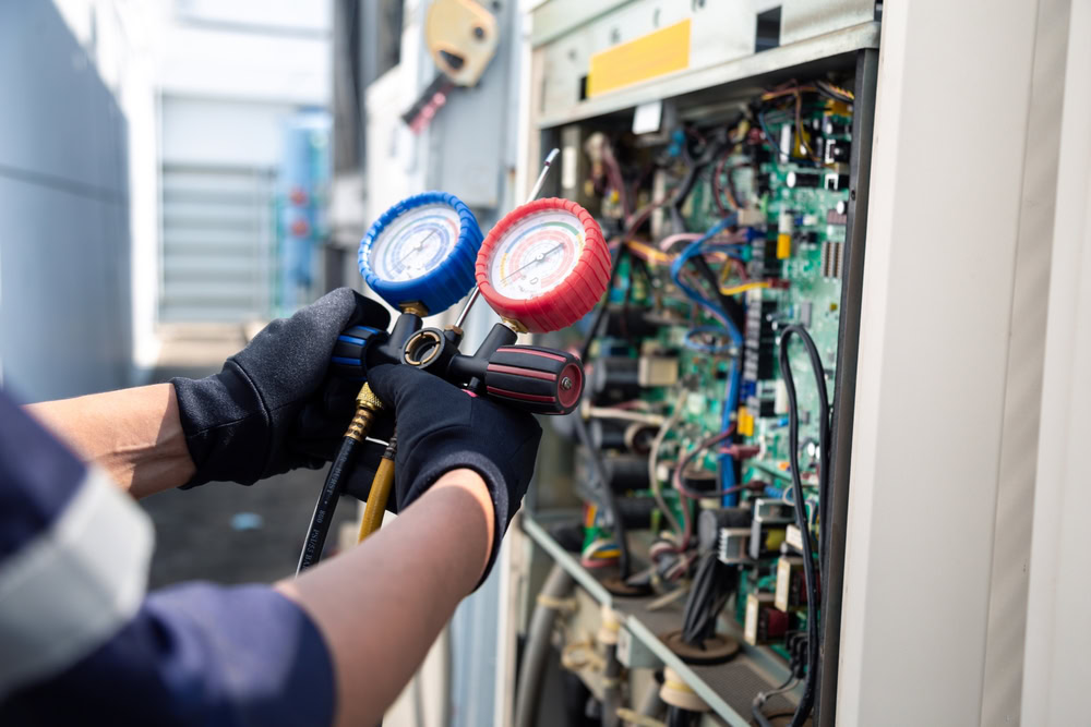 A technician wearing black gloves uses a manifold gauge to check and service the wiring and components inside an open HVAC unit during an emergency HVAC service in Queens, NY. Various colorful wires and circuit boards are visible inside the unit.