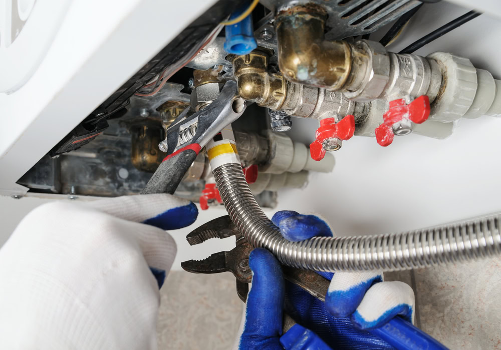 A close-up of a plumber’s gloved hands using tools to tighten fittings on a gas boiler, working with metal pipes and valves—ideal for emergency HVAC service in Long Island or Queens, NY.