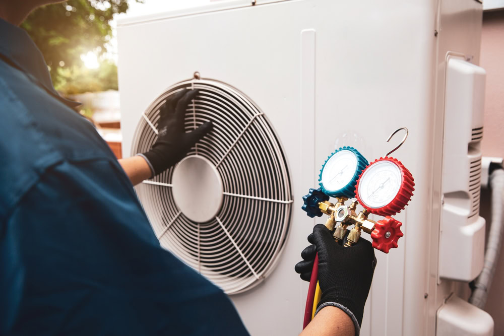 A technician wearing gloves uses a manifold gauge to check or service an outdoor air conditioning unit in Queens, NY, with one hand on the unit's fan.
