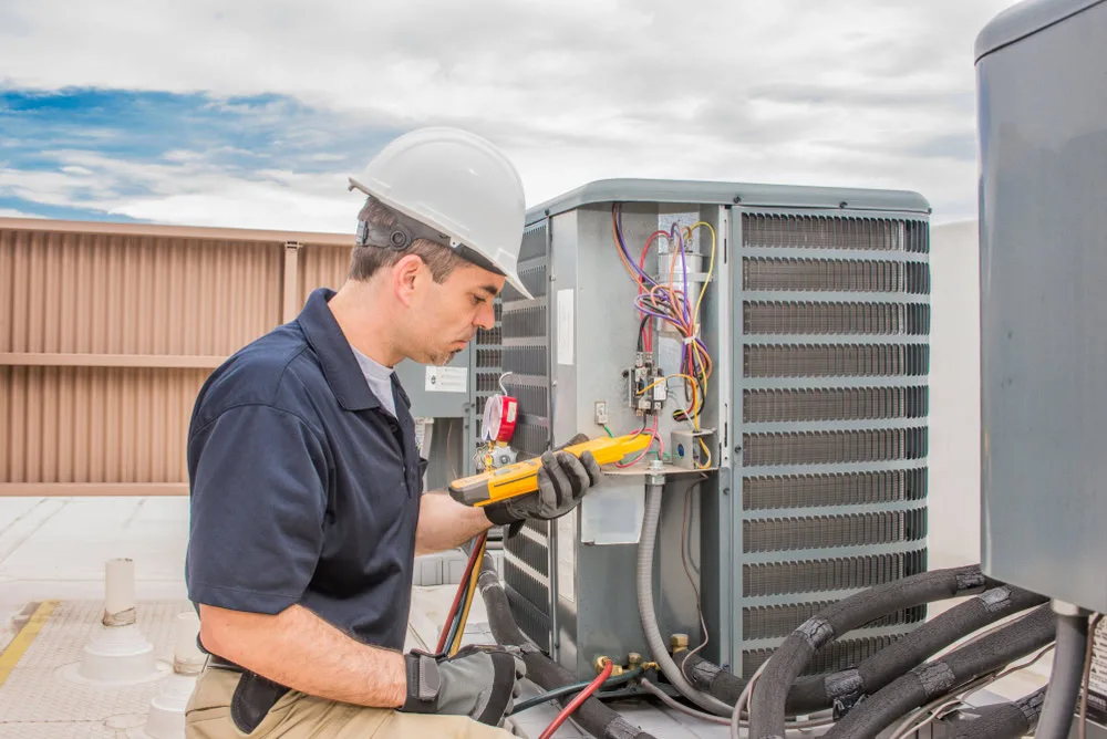 A technician wearing a hard hat and gloves uses a multimeter to inspect rooftop air conditioning wiring, ready to provide emergency HVAC service in Long Island, NY or Queens, with tools and equipment visible nearby.