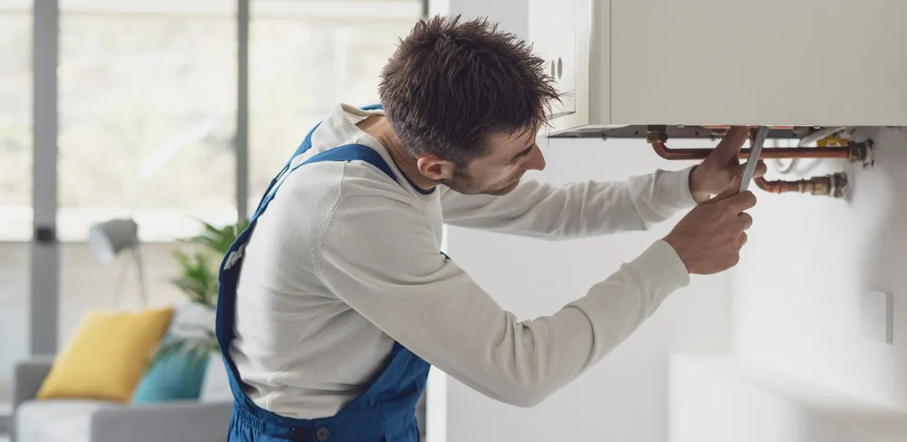 A plumber in blue overalls is repairing or installing pipes under a wall-mounted cabinet in a modern, well-lit living space.