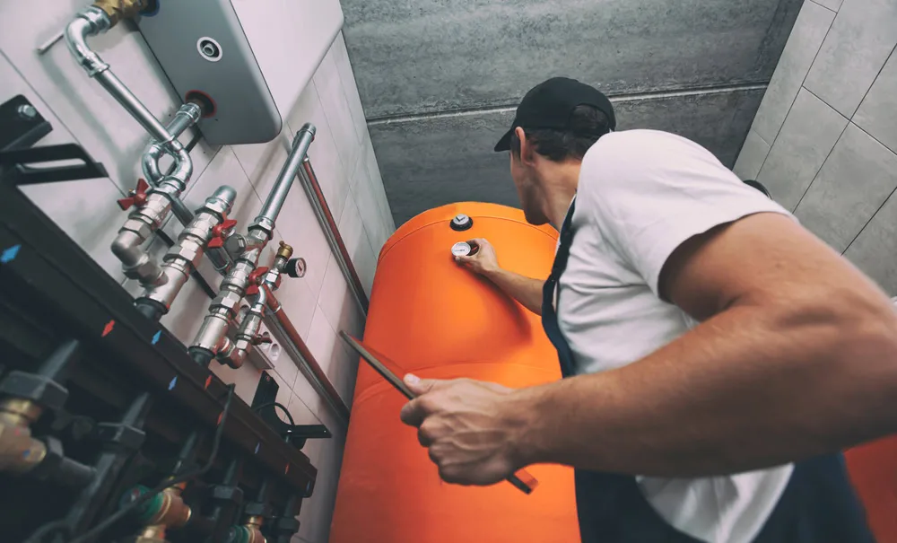 A plumber wearing a cap and overalls checks a gauge on a large orange water tank in a utility room with exposed pipes.