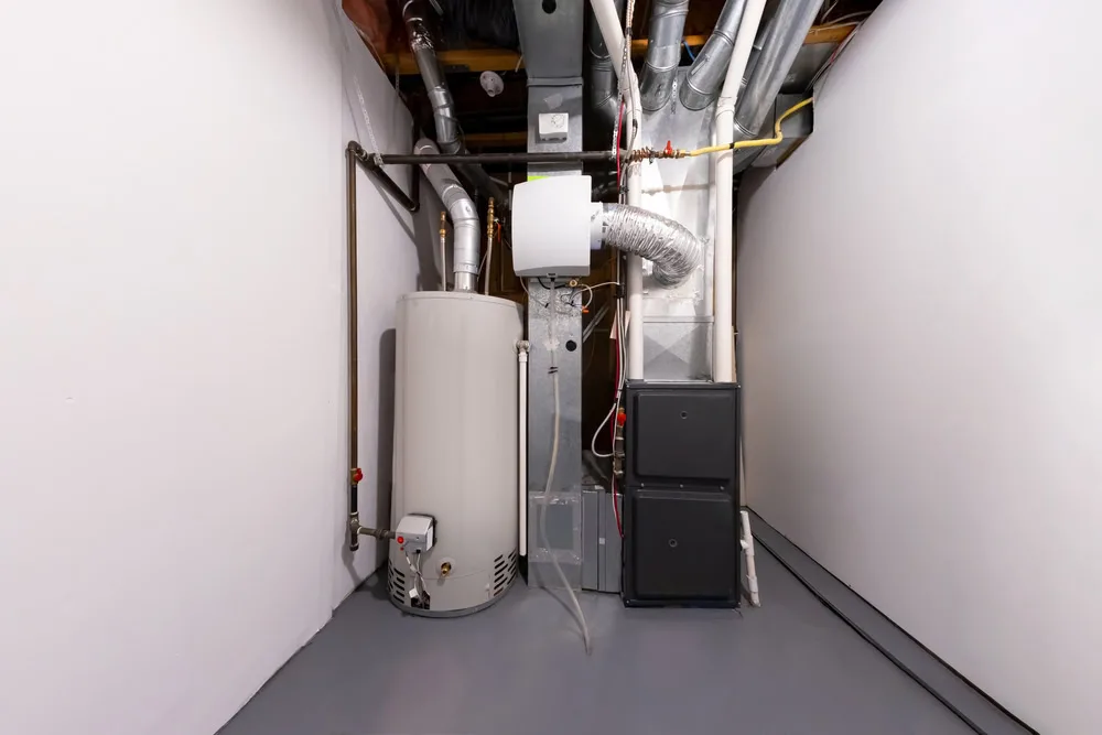 A clean utility room in Queens, NY, features a water heater, furnace, and exposed ductwork against white walls and a gray floor. Pipes and wiring are visible, with neatly installed mechanical systems—ideal for easy emergency HVAC service on Long Island.
