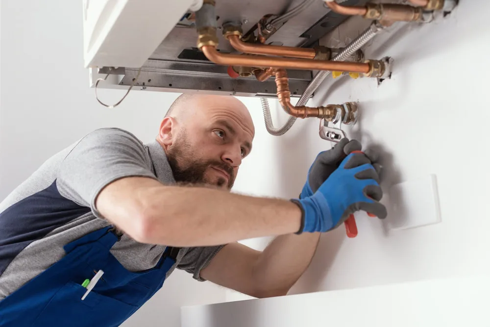 A plumber wearing blue gloves and overalls uses a wrench to fix copper pipes connected to a boiler mounted on a white wall.