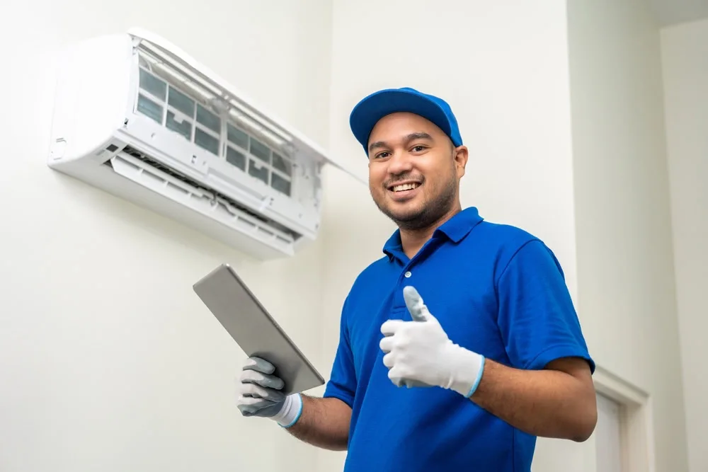A smiling technician in a blue uniform and gloves gives a thumbs up while holding a tablet, standing next to an open wall-mounted air conditioner—ready to provide emergency HVAC service Long Island or Queens, NY residents can trust.