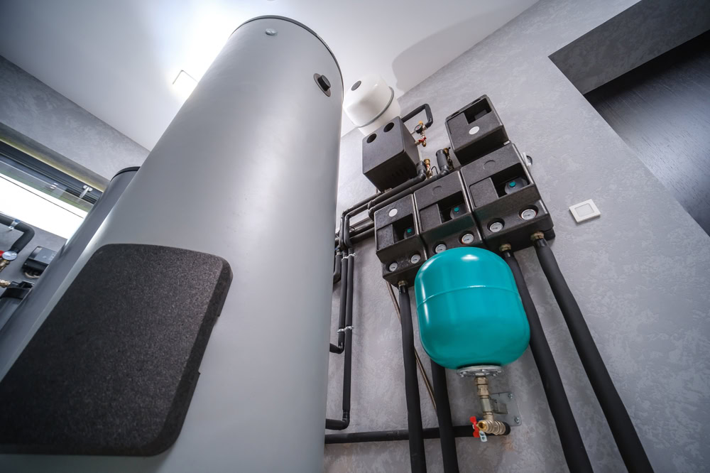 Close-up view of a modern boiler room with a large cylindrical hot water tank, insulated pipes, control units, and a prominent green expansion vessel mounted on a gray wall.