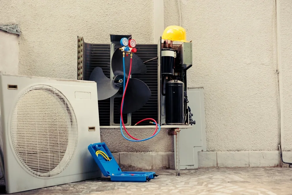 An outdoor air conditioning unit being serviced for emergency HVAC service in Queens, with its cover open to reveal internal components, attached gauges, a yellow hard hat on top, and a blue tool case on the ground nearby against a beige wall.