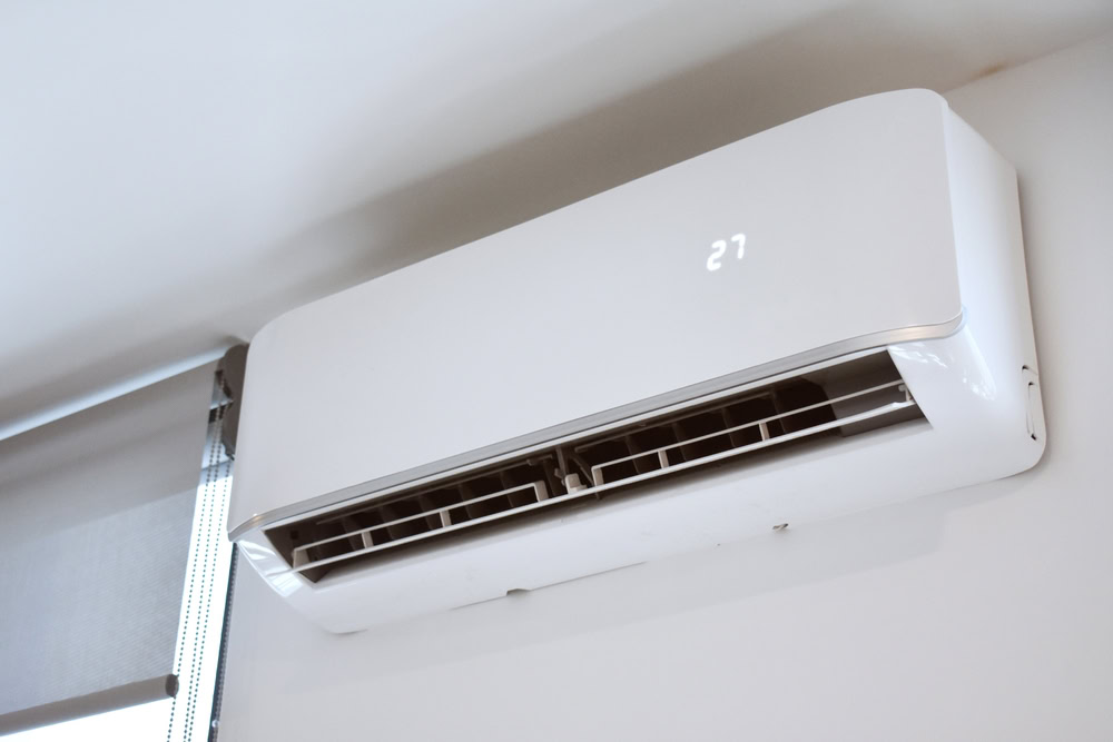A white wall-mounted air conditioner displays "27" on its panel, installed near a window with the blind down. For prompt repairs or emergency HVAC service in Queens, NY or Long Island, trust local experts to keep your space comfortable.