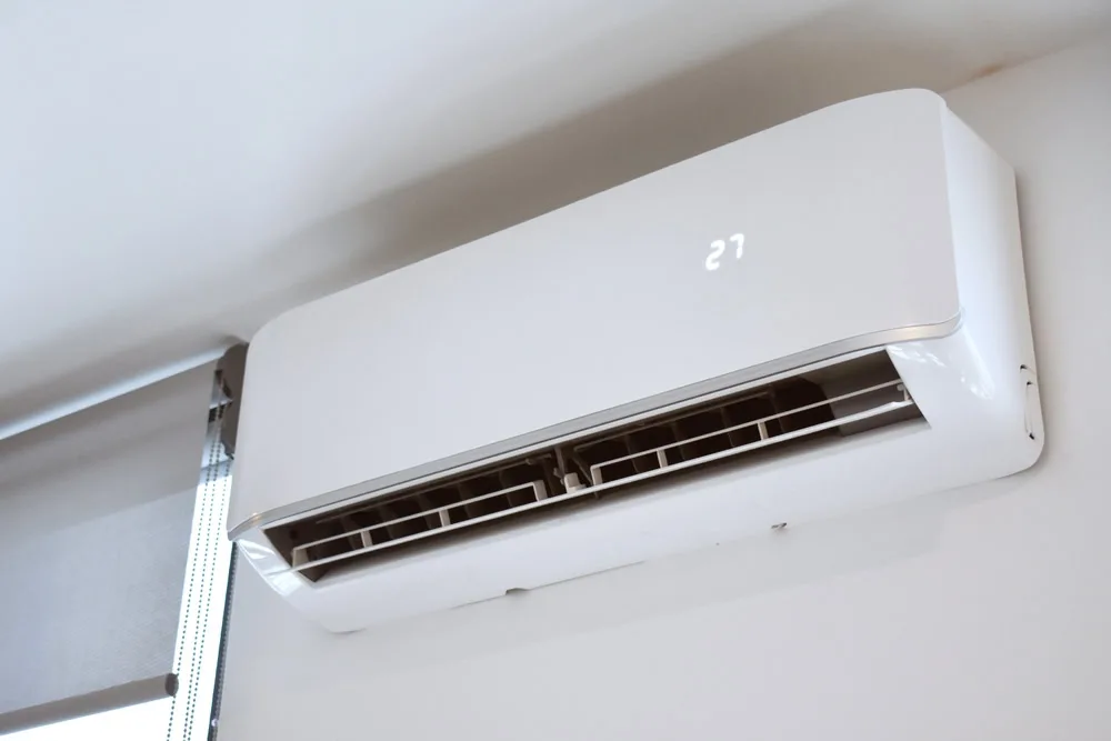 A white wall-mounted air conditioner displays "27" on its panel, installed near a window with the blind down. For prompt repairs or emergency HVAC service in Queens, NY or Long Island, trust local experts to keep your space comfortable.