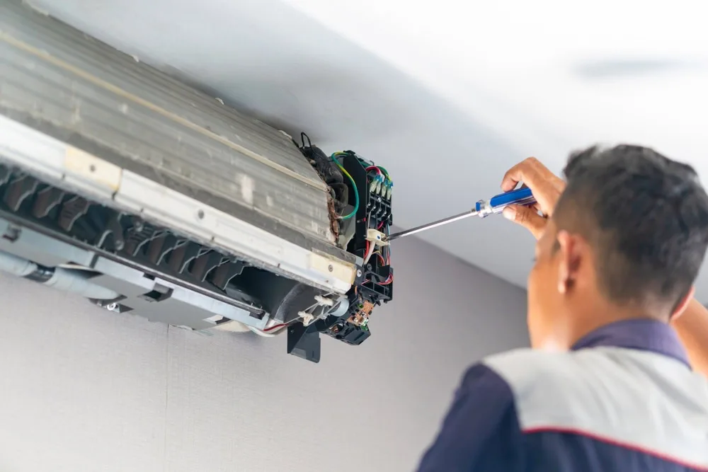 A technician provides emergency HVAC service Long Island, using a screwdriver to repair or maintain the internal components of a wall-mounted air conditioning unit.