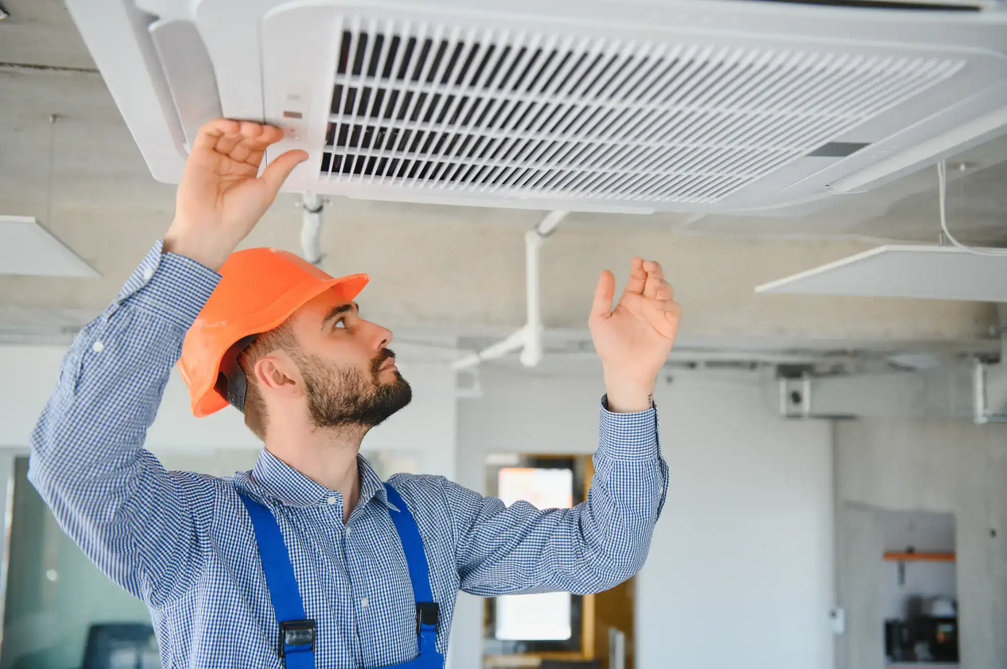 A worker in a checkered shirt and orange hard hat inspects or installs a ceiling air conditioning unit in a modern indoor space.