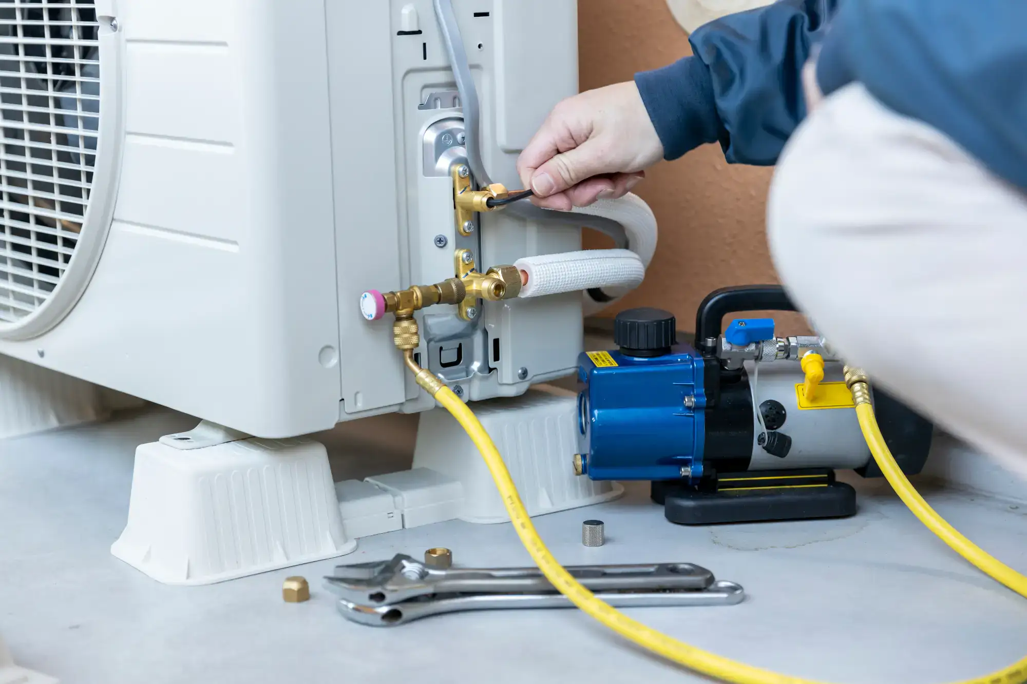 A person installs or maintains an air conditioning unit, using a wrench to adjust connectors attached to yellow hoses and a blue vacuum pump on the ground nearby.