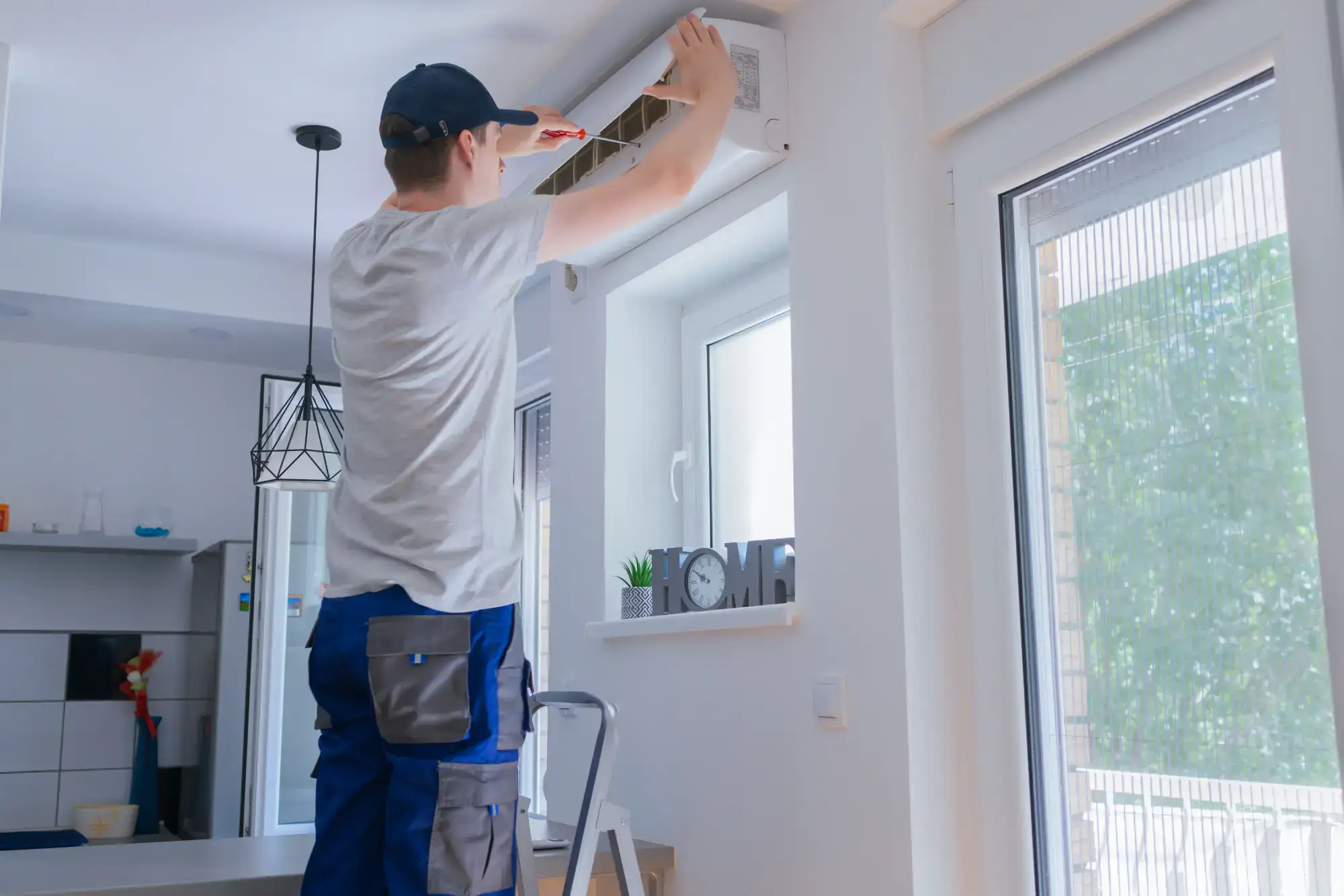 A technician in a cap and work clothes stands on a step ladder, servicing an air conditioning unit mounted high on a white wall in a bright, modern room with large windows.