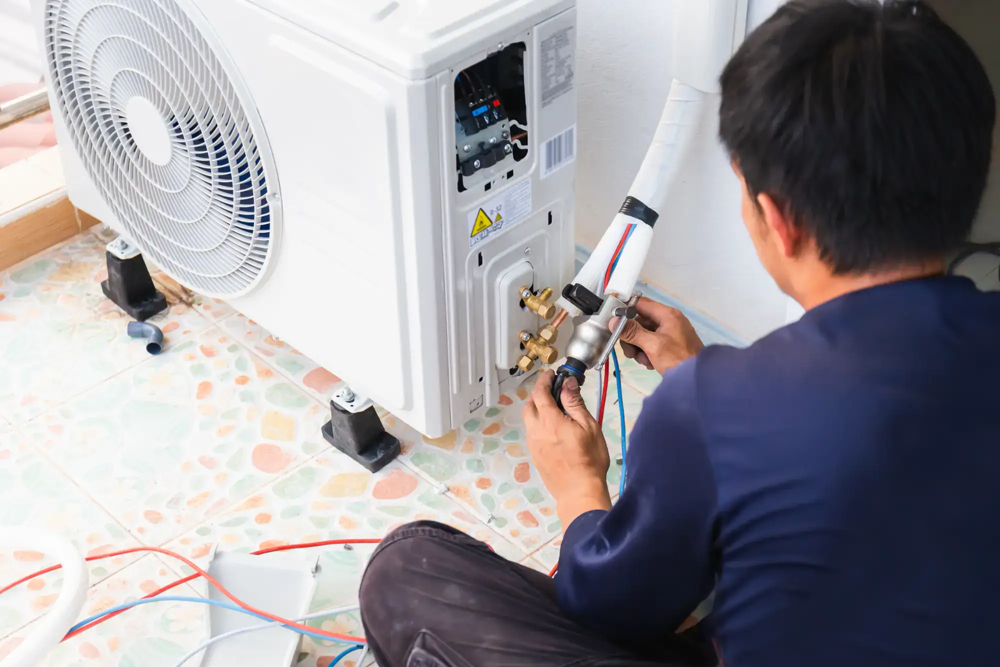 A technician kneels on a tiled floor while installing or repairing an air conditioning unit, connecting wires and pipes to the outdoor unit.