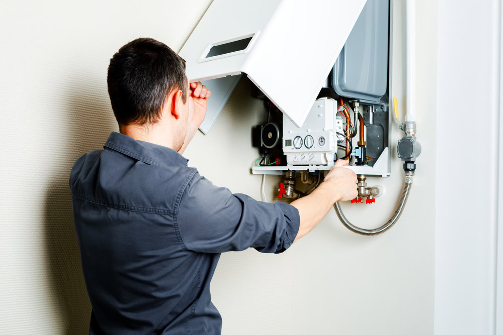 Technician inspecting and repairing a wall‑mounted boiler or gas water heater with the front panel open during home heating system maintenance.